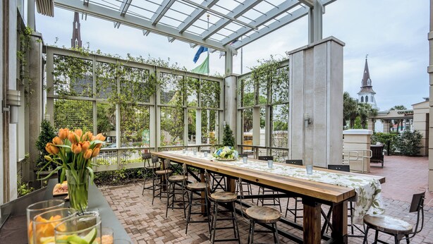 An outdoor dining area with a long wooden table and multiple stools, surrounded by glass walls and a brick floor. - Courtyard Charleston Historic District (Charleston)