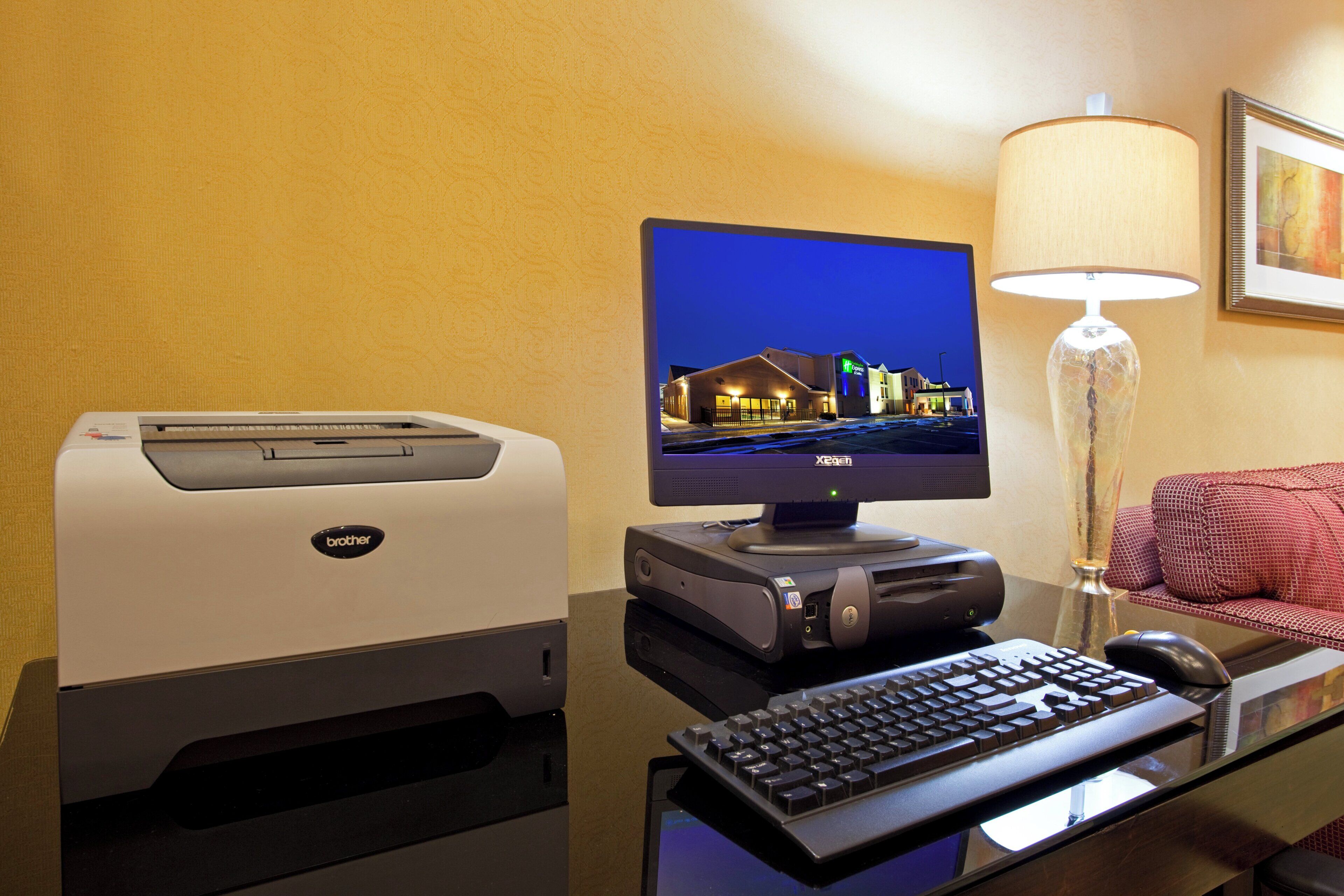 Habitación de hotel con un ordenador de escritorio, una impresora, un teclado, una lámpara y un cuadro en la pared.
