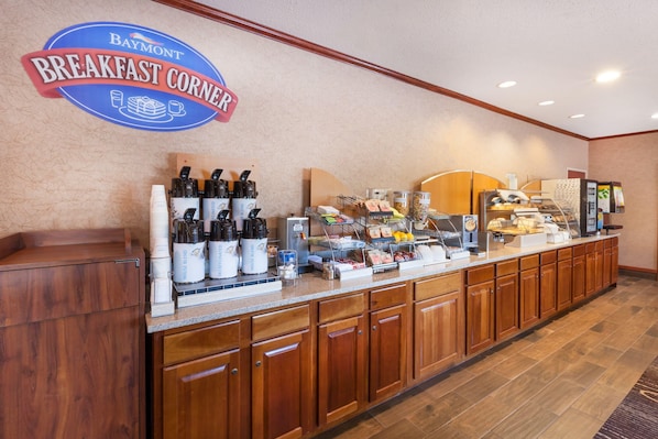 A breakfast buffet counter with a sign reading 'Baymont Breakfast Corner,' various food items, and coffee machines.