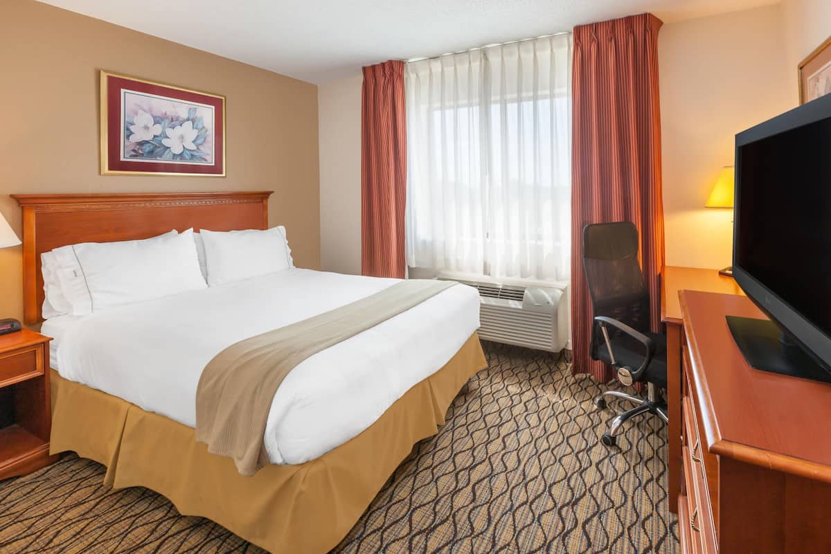 A hotel room with a large bed, a desk with a television, a chair, and a window with curtains.