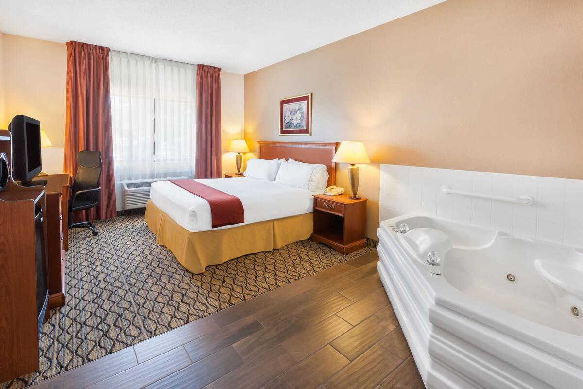 A hotel room with a bed, a desk with a computer, a desk lamp, a telephone, a TV, a window with curtains, a painting on the wall, and a bathtub.