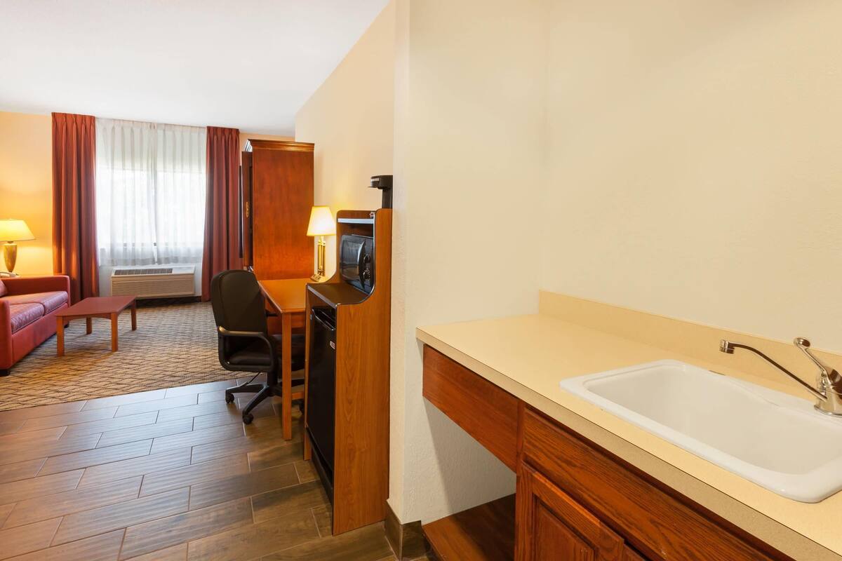 A hotel room with a wooden desk, a chair, a sofa, a small table, a lamp, a TV, and a sink.