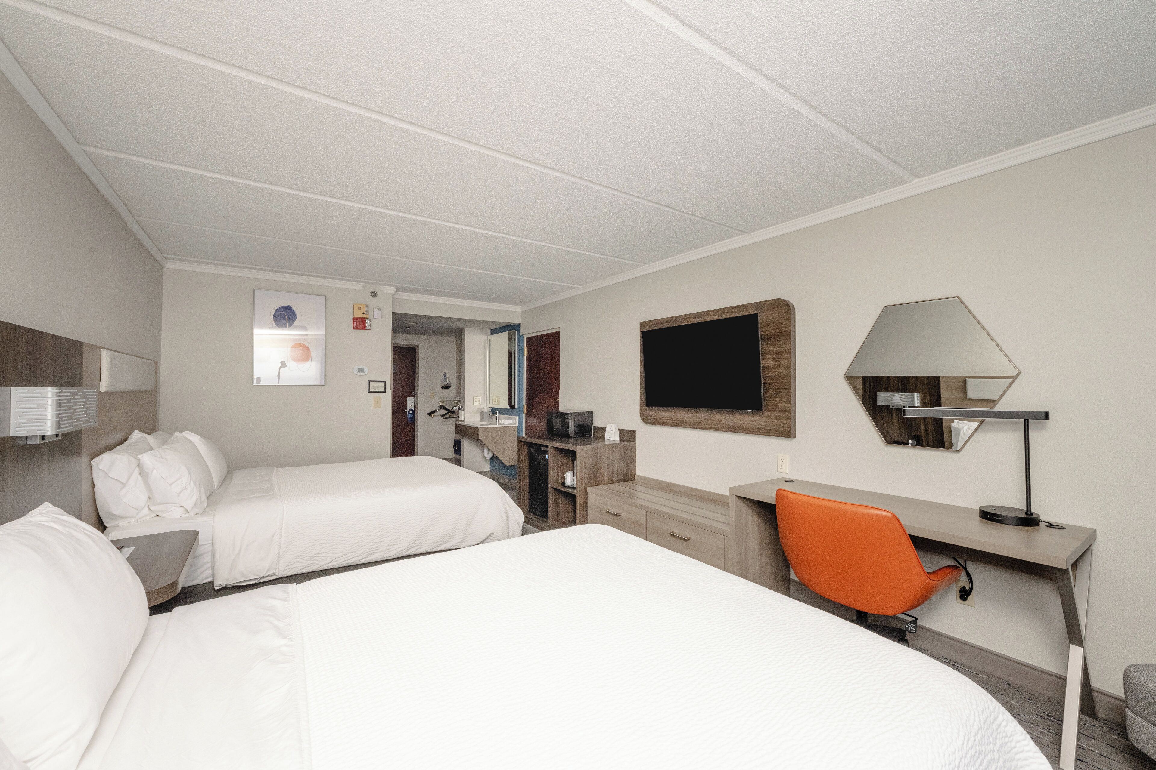 Una habitación de hotel con dos camas, un escritorio, una silla naranja, un televisor y un espejo.