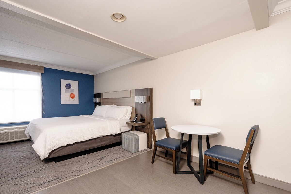 A hotel room with a bed, a desk with a chair, a small table, and a wall-mounted lamp.