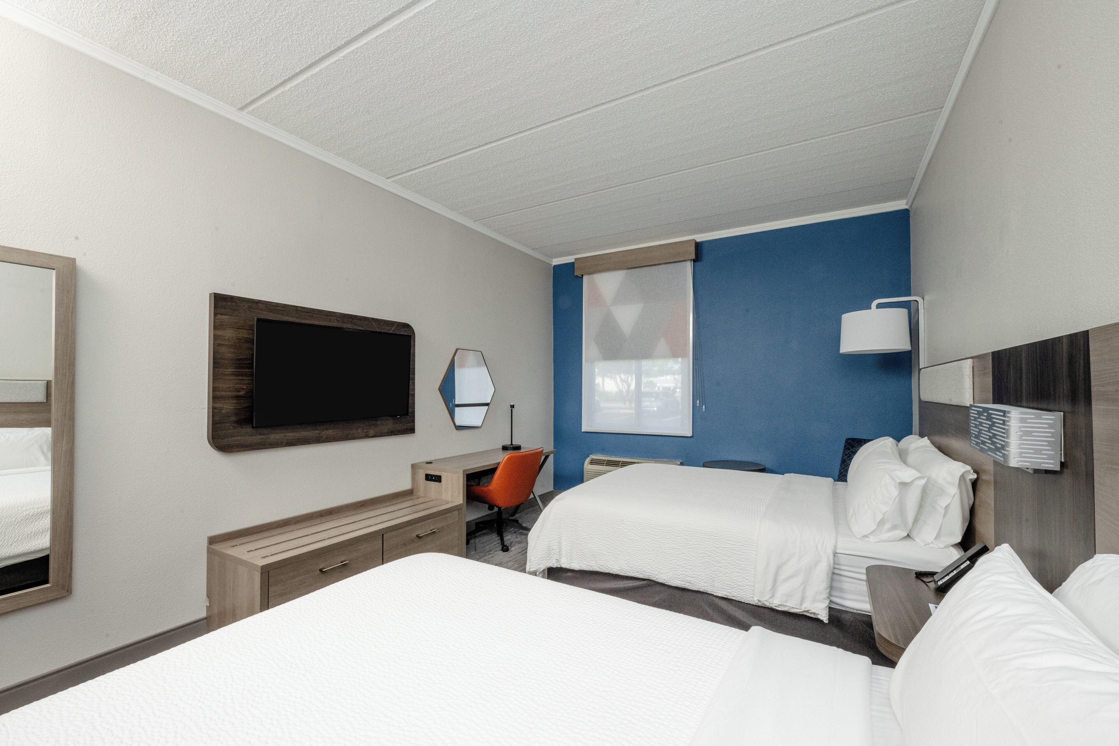 A hotel room with two beds, a desk, an orange chair, a TV, and a window with blinds.