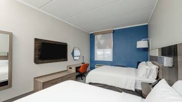 A hotel room with two beds, a desk, an orange chair, a TV, and a window with blinds.