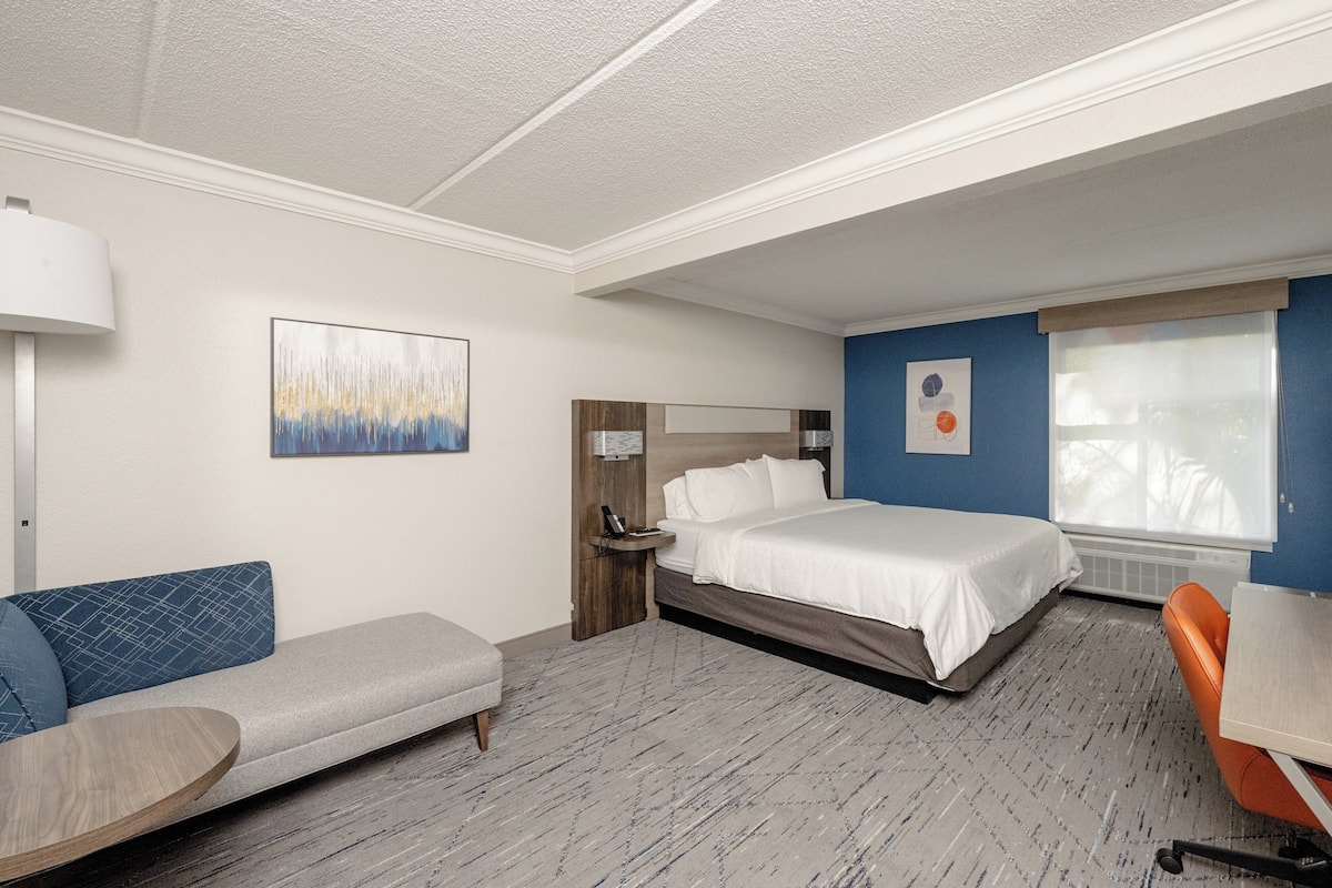 A hotel room with a bed, a sofa, a desk, a lamp, and artwork on the walls.