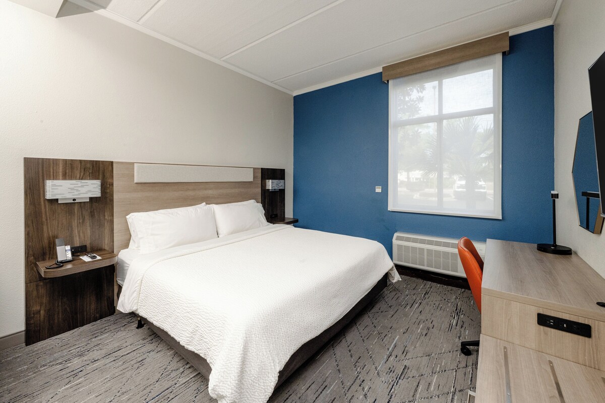 A hotel room with a bed, desk, chair, nightstand, and a window with blinds.