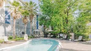 Una piscina con sillas de descanso y sombrillas, rodeada de palmeras y un edificio con amplias ventanas.