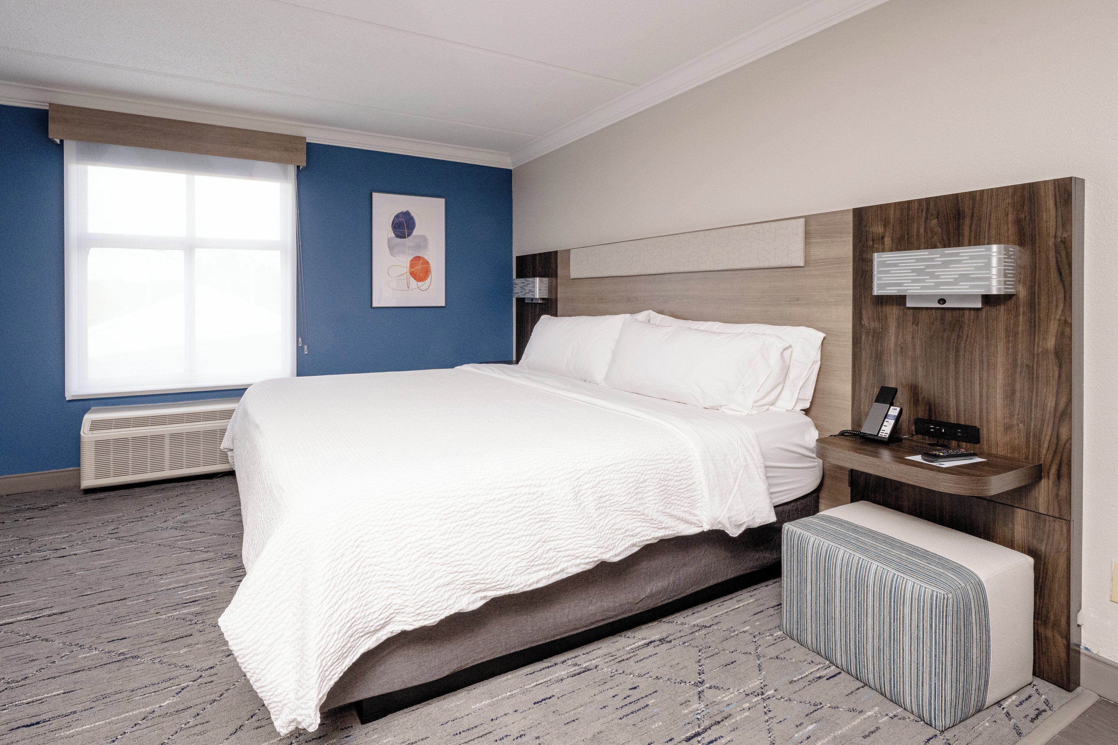 A modern hotel room with a large bed, a bedside table with a phone, a bench, and a wall-mounted light fixture.