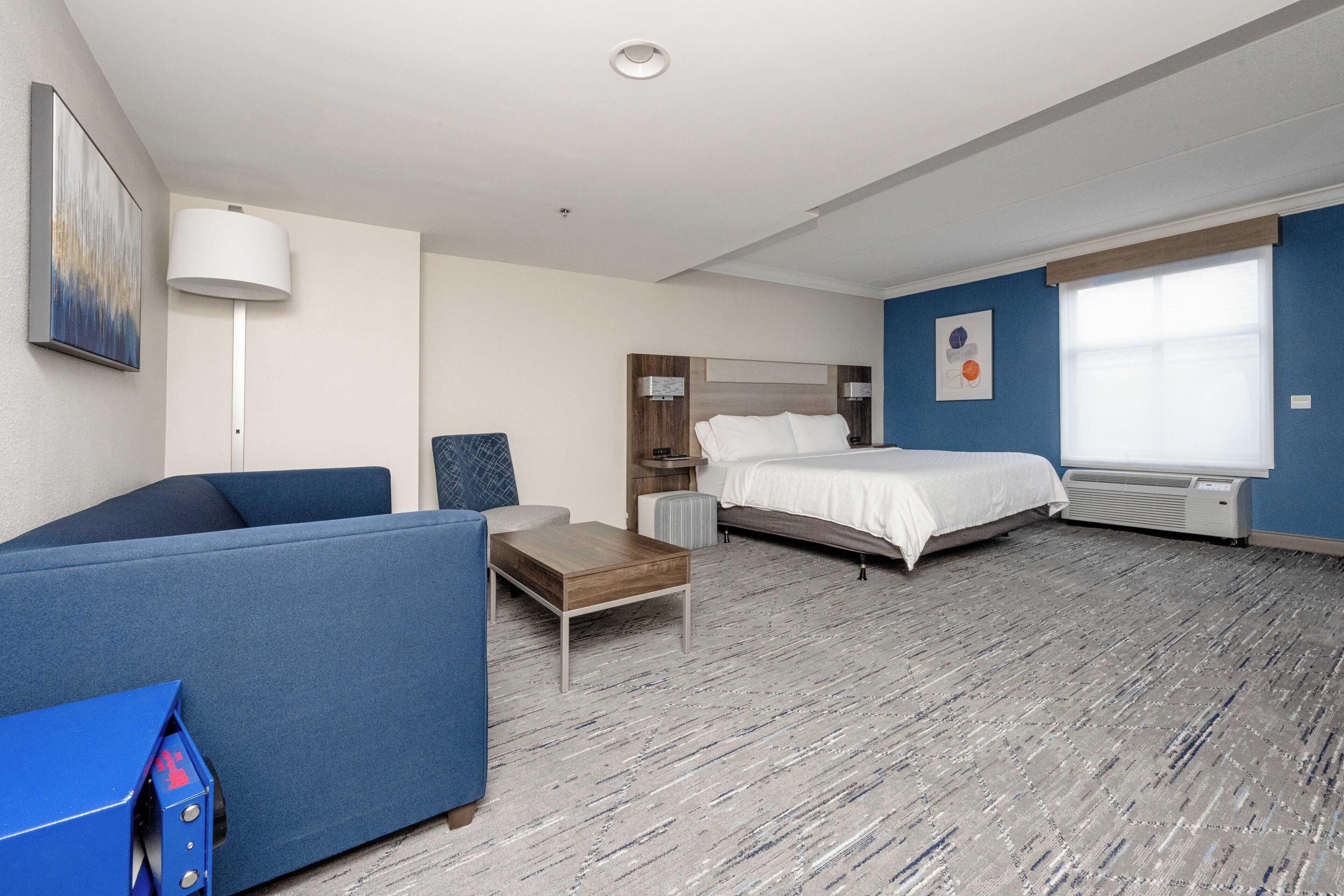 A hotel room with a bed, a blue sofa, a small table, a lamp, and a window.