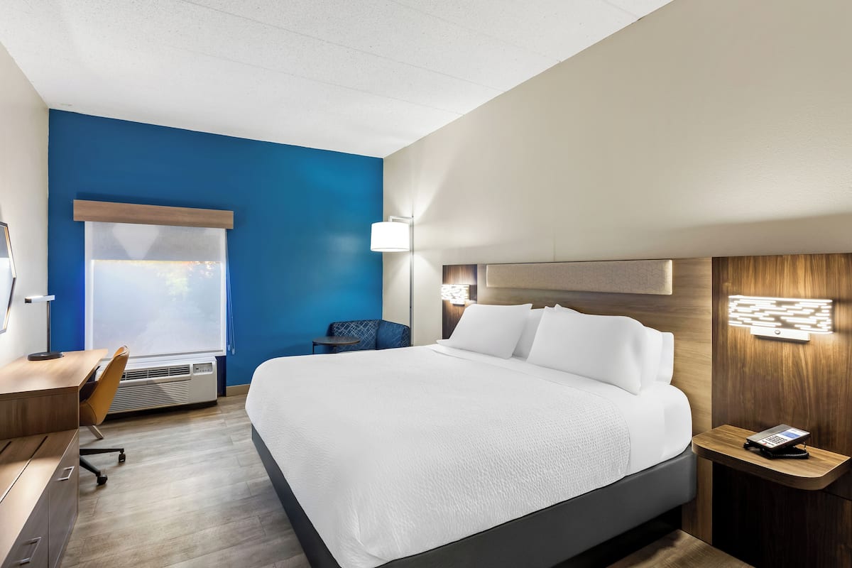 Une chambre d’hôtel avec un grand lit, un bureau, une chaise, une table de chevet avec un téléphone et une fenêtre avec des stores.