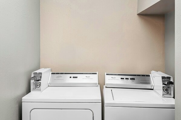 Two white front-loading washing machines in a laundry area with a plain wall and a corner cabinet.