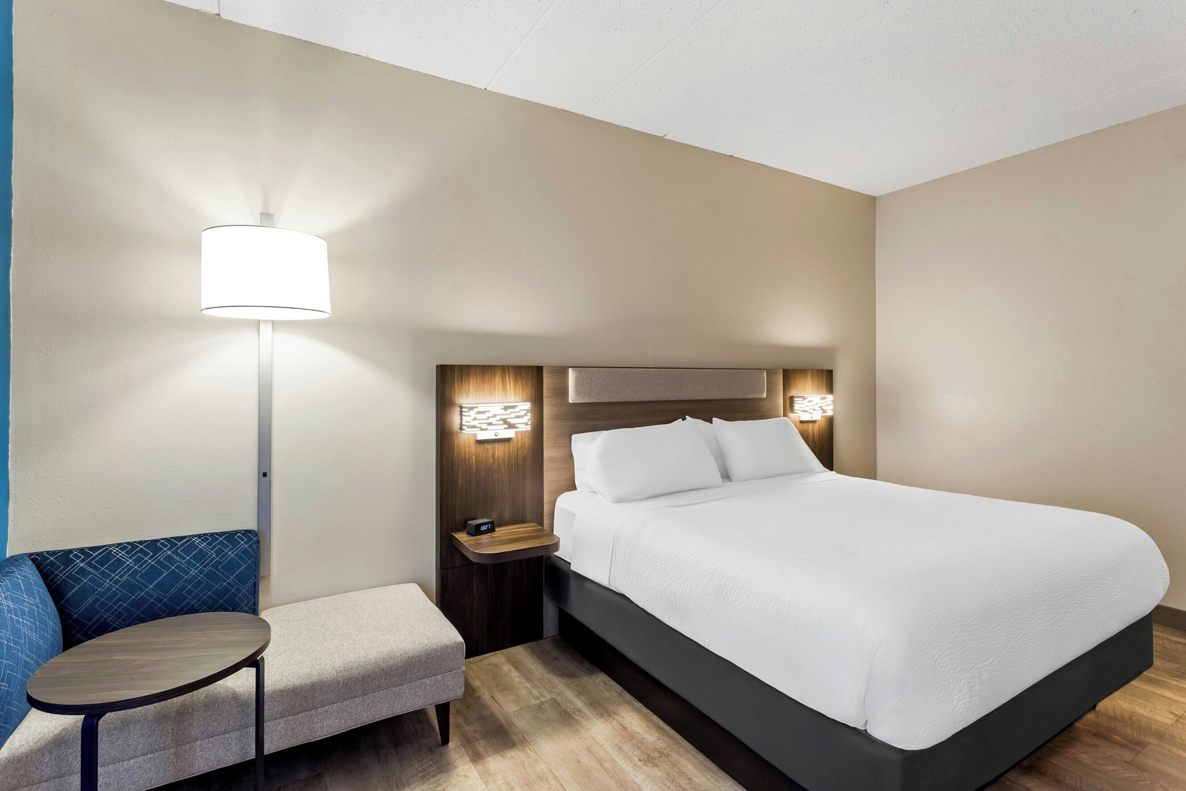 Una habitación de hotel con cama, mesita de noche, lámpara, silla y una pequeña mesa.