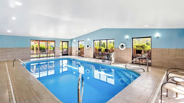 A swimming pool with a tiled floor, surrounded by chairs and tables, and large windows offering views of greenery.