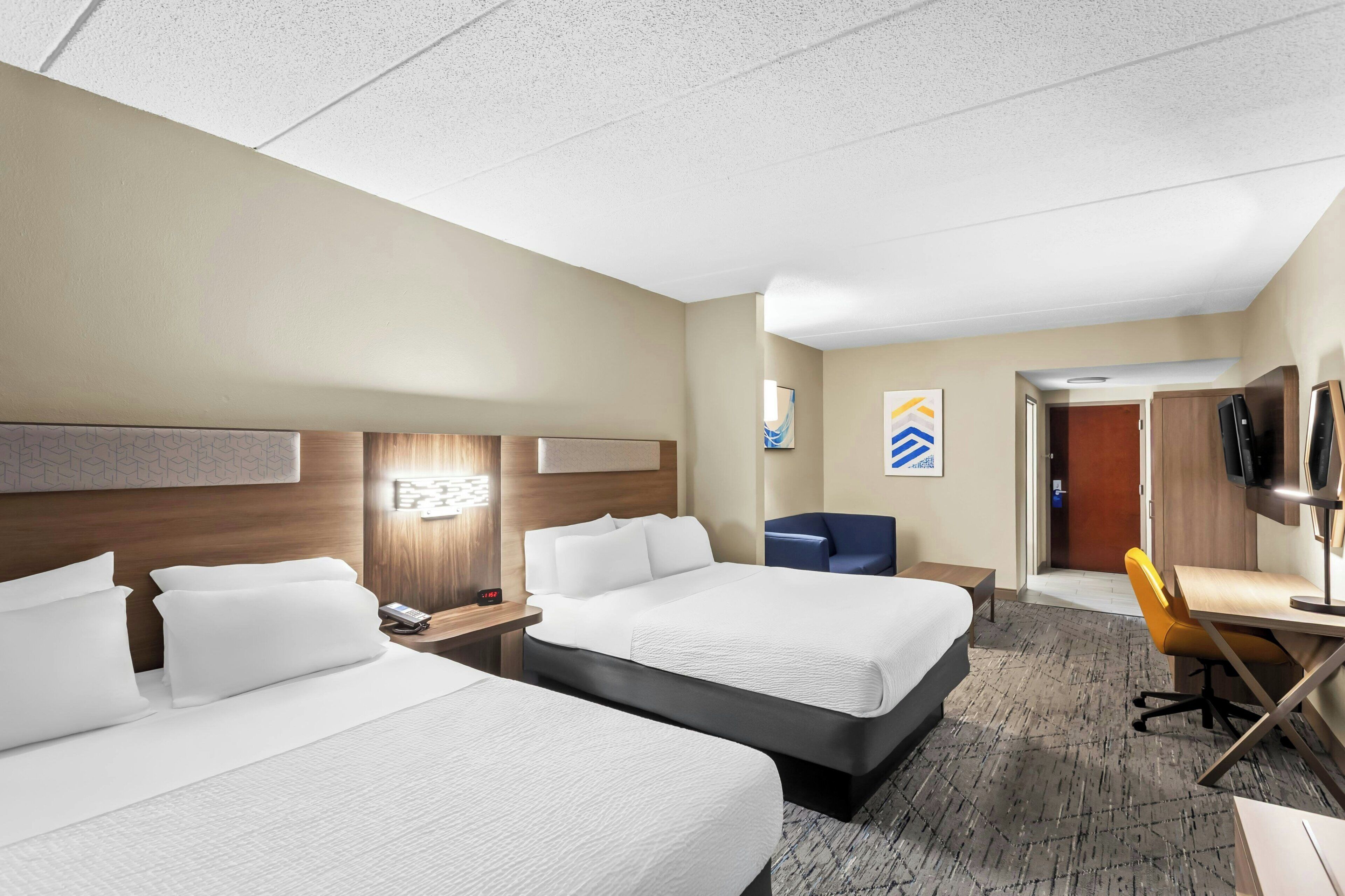 Quarto de hotel com duas camas, uma escrivaninha, uma cadeira, uma televisão e uma porta que dá para outro cômodo.