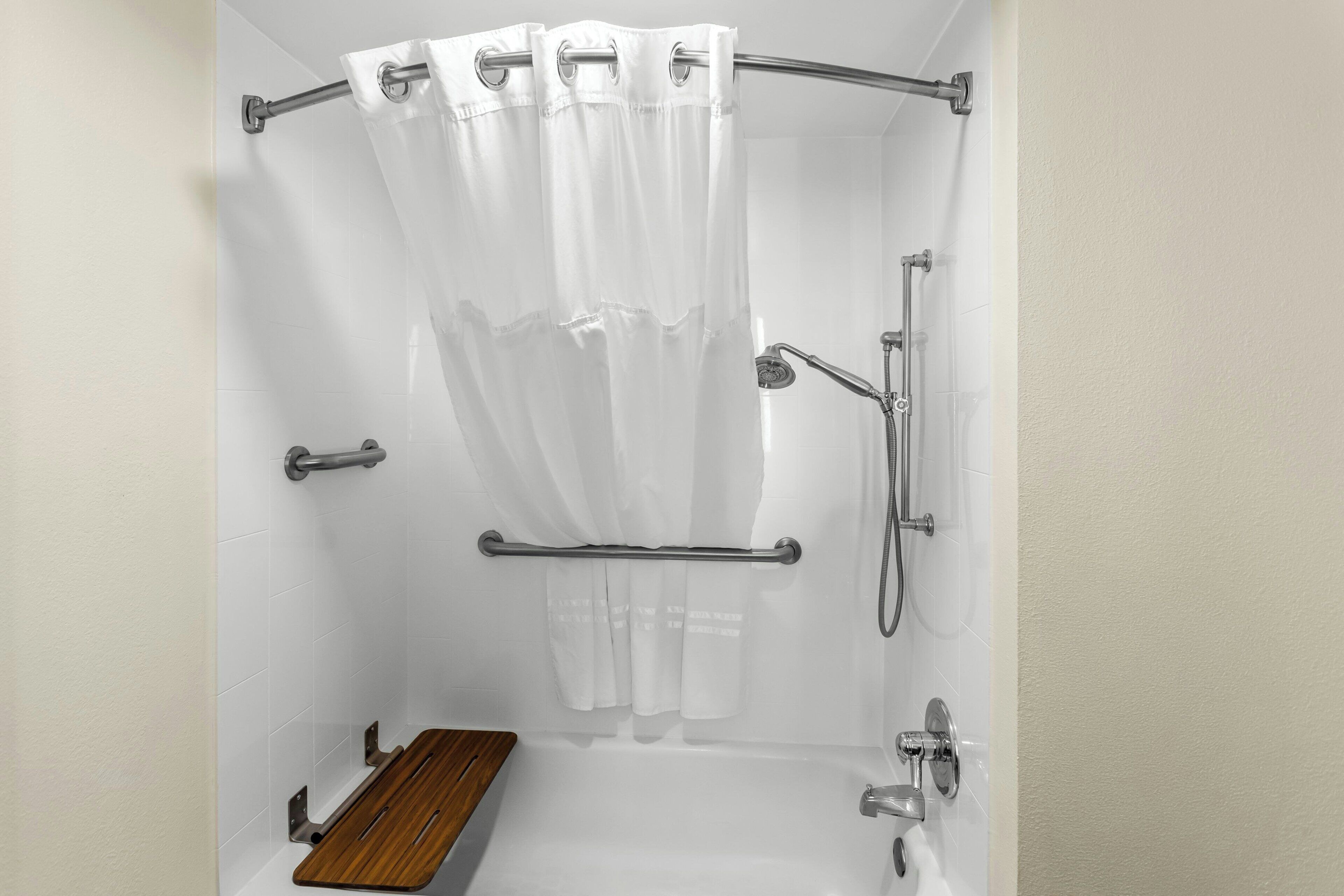 A shower with a white curtain, grab bars, a handheld showerhead, and a wooden bench.