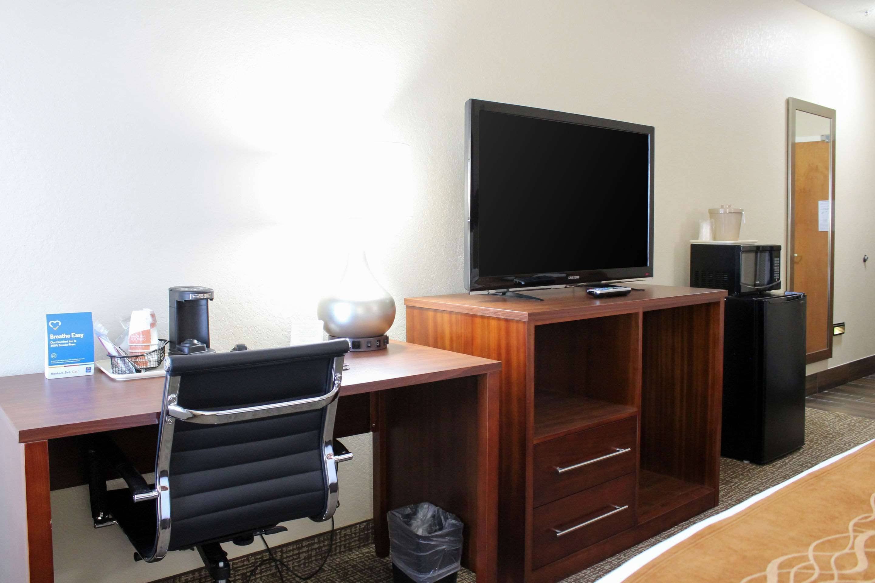 A hotel room with a wooden desk, a black office chair, a flat-screen TV, a microwave, a trash bin, and a coffee maker.