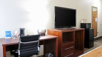 A hotel room with a wooden desk, a black office chair, a flat-screen TV, a microwave, a trash bin, and a coffee maker.