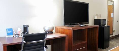 A hotel room with a wooden desk, a black office chair, a flat-screen TV, a microwave, a trash bin, and a coffee maker.