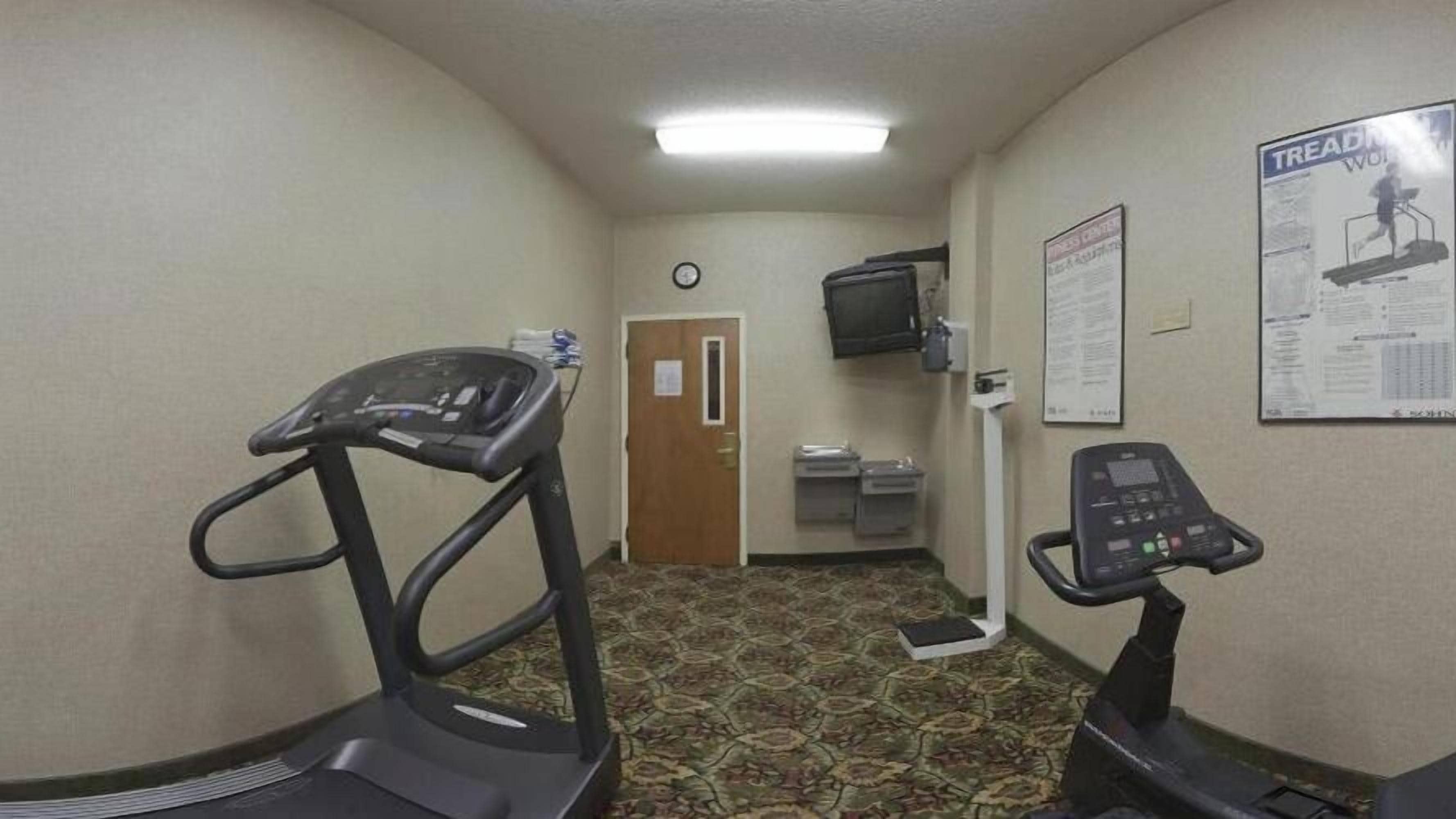 A hallway with a treadmill, a stationary bike, a clock, a television, and a poster on the wall.