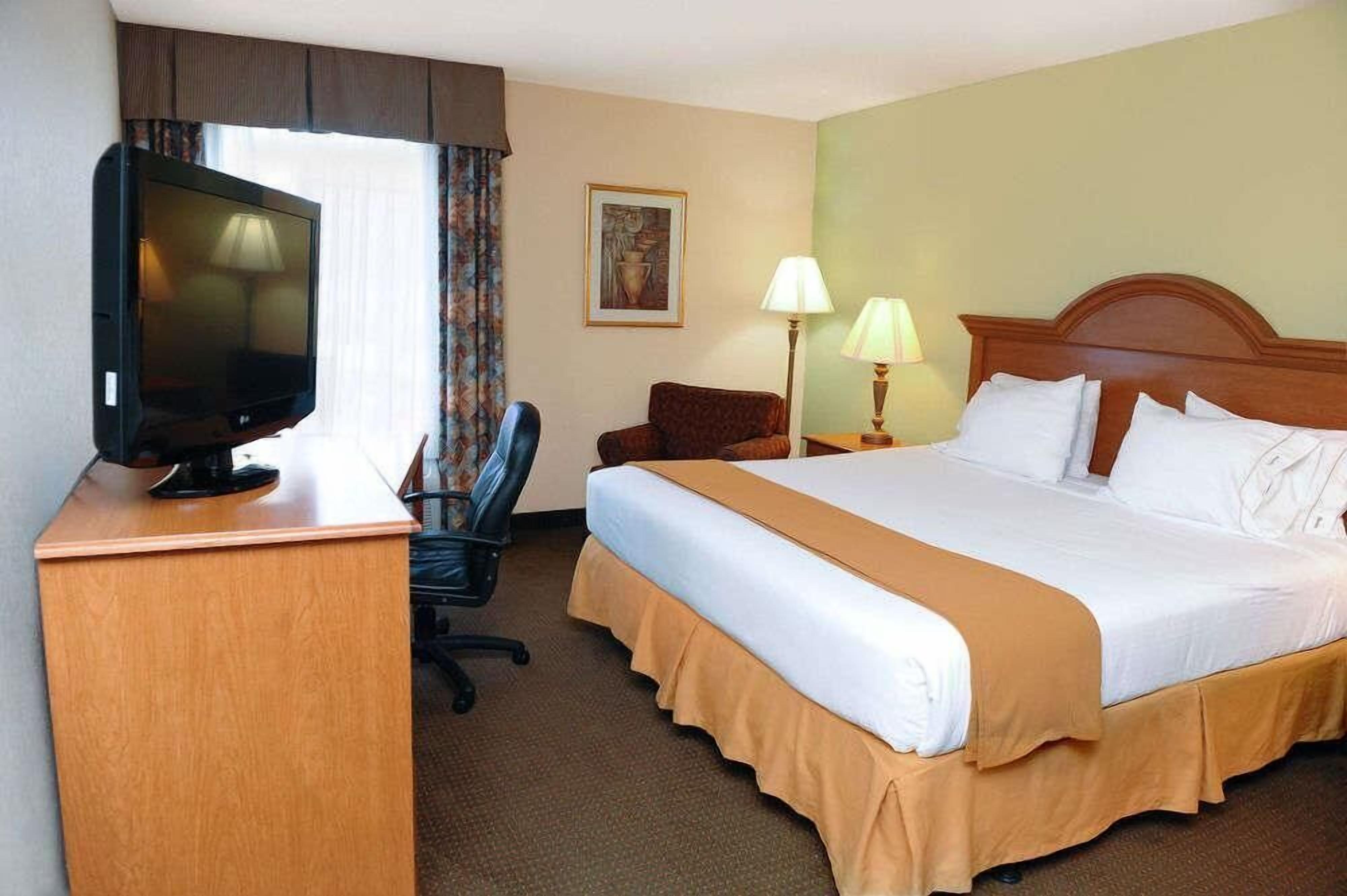 A hotel room with a bed, a desk with a television, a chair, a lamp, and a painting on the wall.