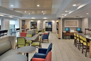 A modern hotel dining area with multiple seating options including tables and booths, a kitchen area in the background, and a patterned wall decoration. - Holiday Inn Express Raleigh Durham Airport by IHG (Morrisville)