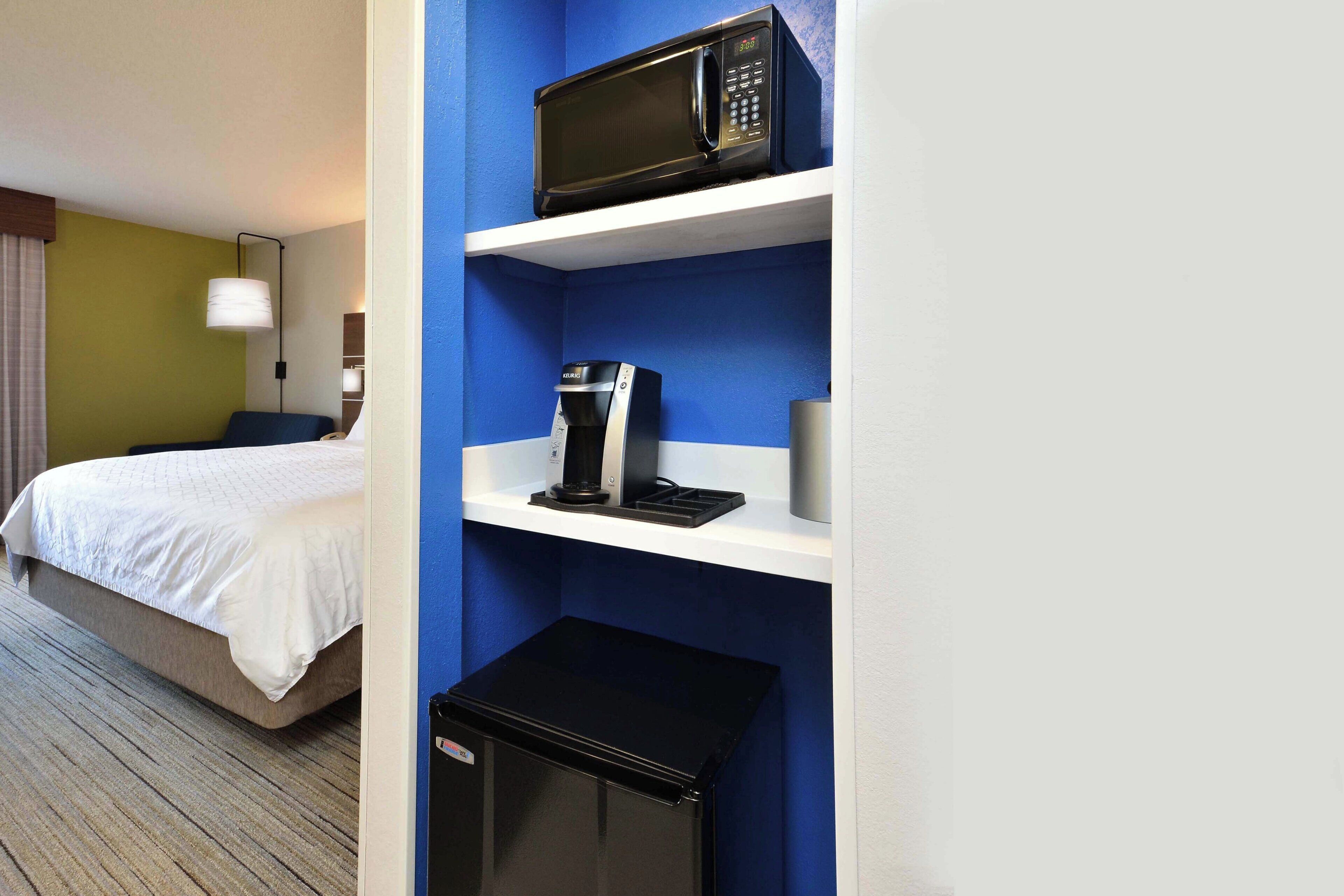 Quarto de hotel com cama, micro-ondas, cafeteira e pequena geladeira.