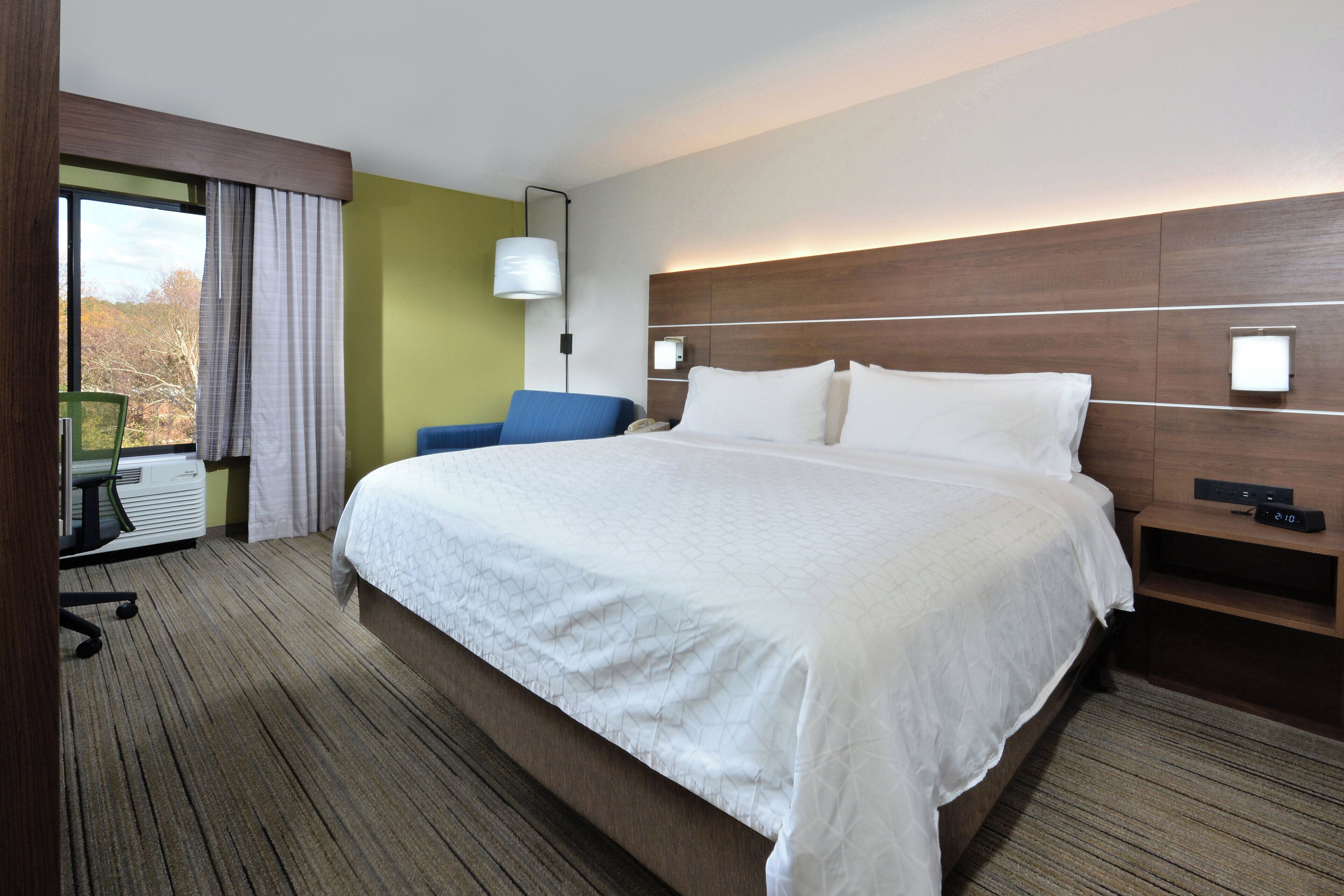 Quarto de hotel com uma cama grande, uma mesa de escritório, uma cadeira, abajur e janela com cortinas.