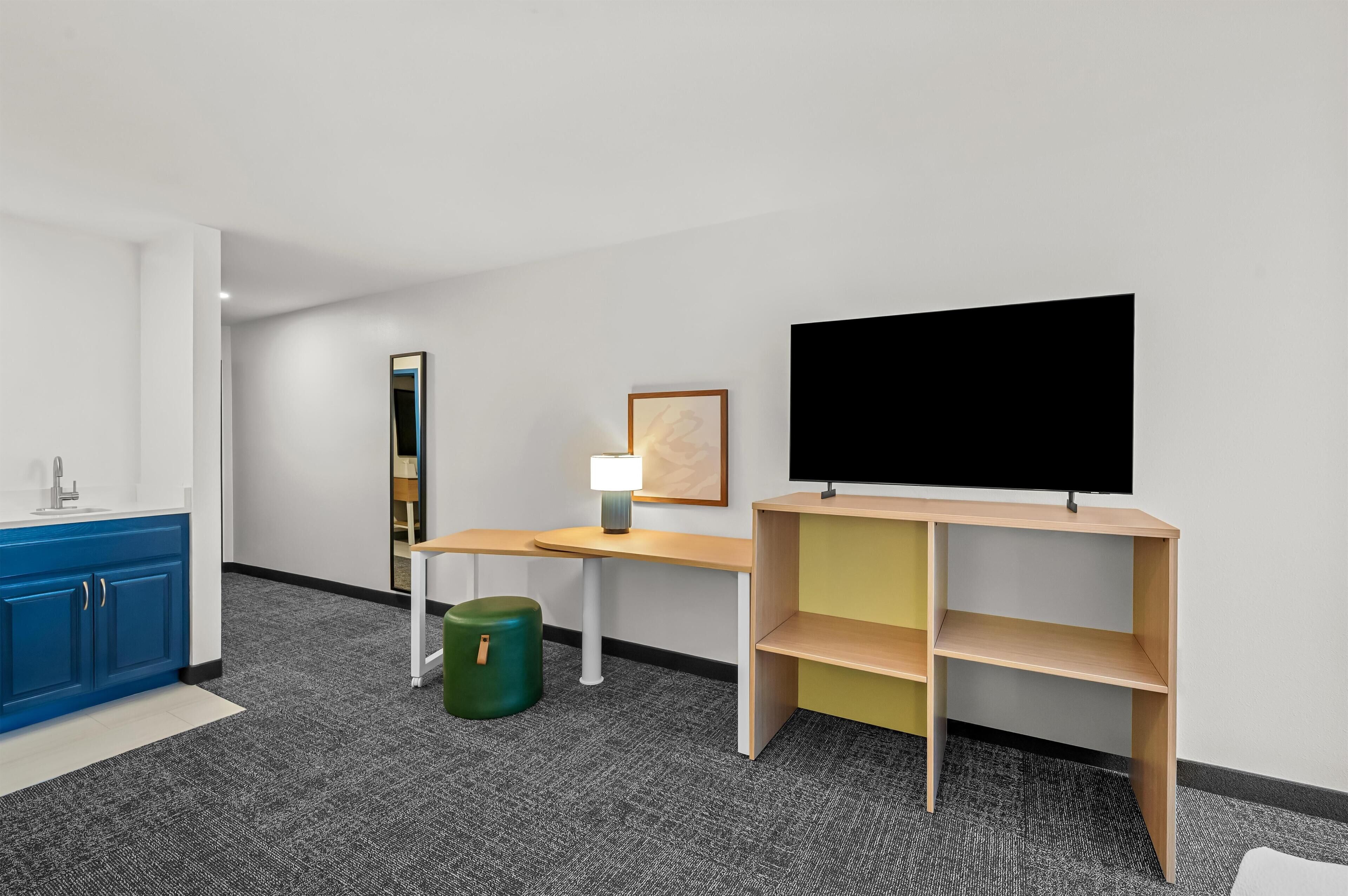 Une chambre d’hôtel moderne équipée d’une kitchenette, d’un bureau, d’une télévision, d’une lampe et d’une poubelle verte.