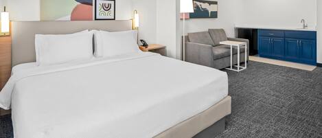A hotel room with a large bed, a desk, a chair, a small table, a blue cabinet, and a wall-mounted lamp.
