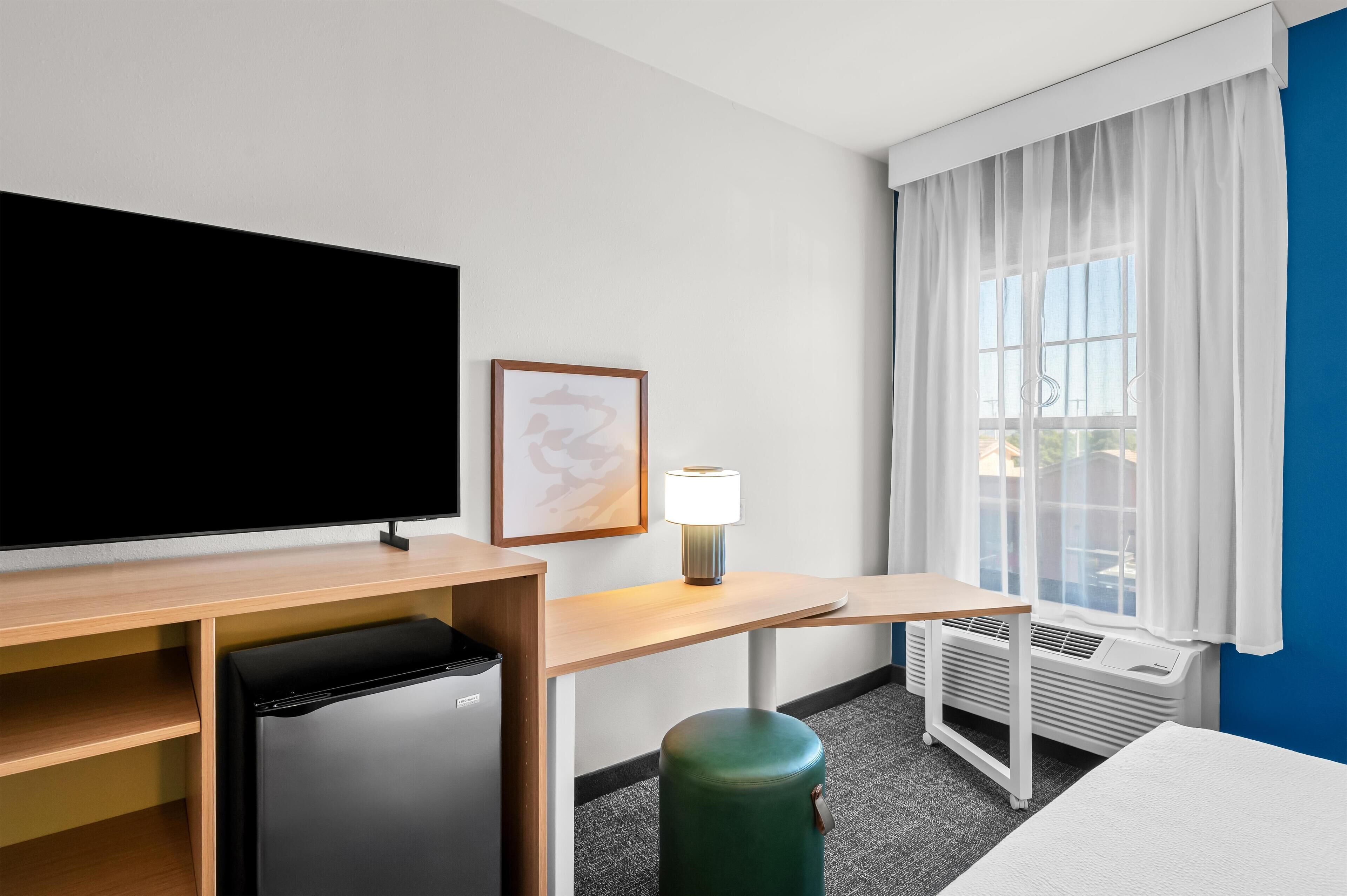 Hotelkamer met flatscreen televisie, kleine koelkast, bureau met lamp, raam met witte gordijnen en radiator.