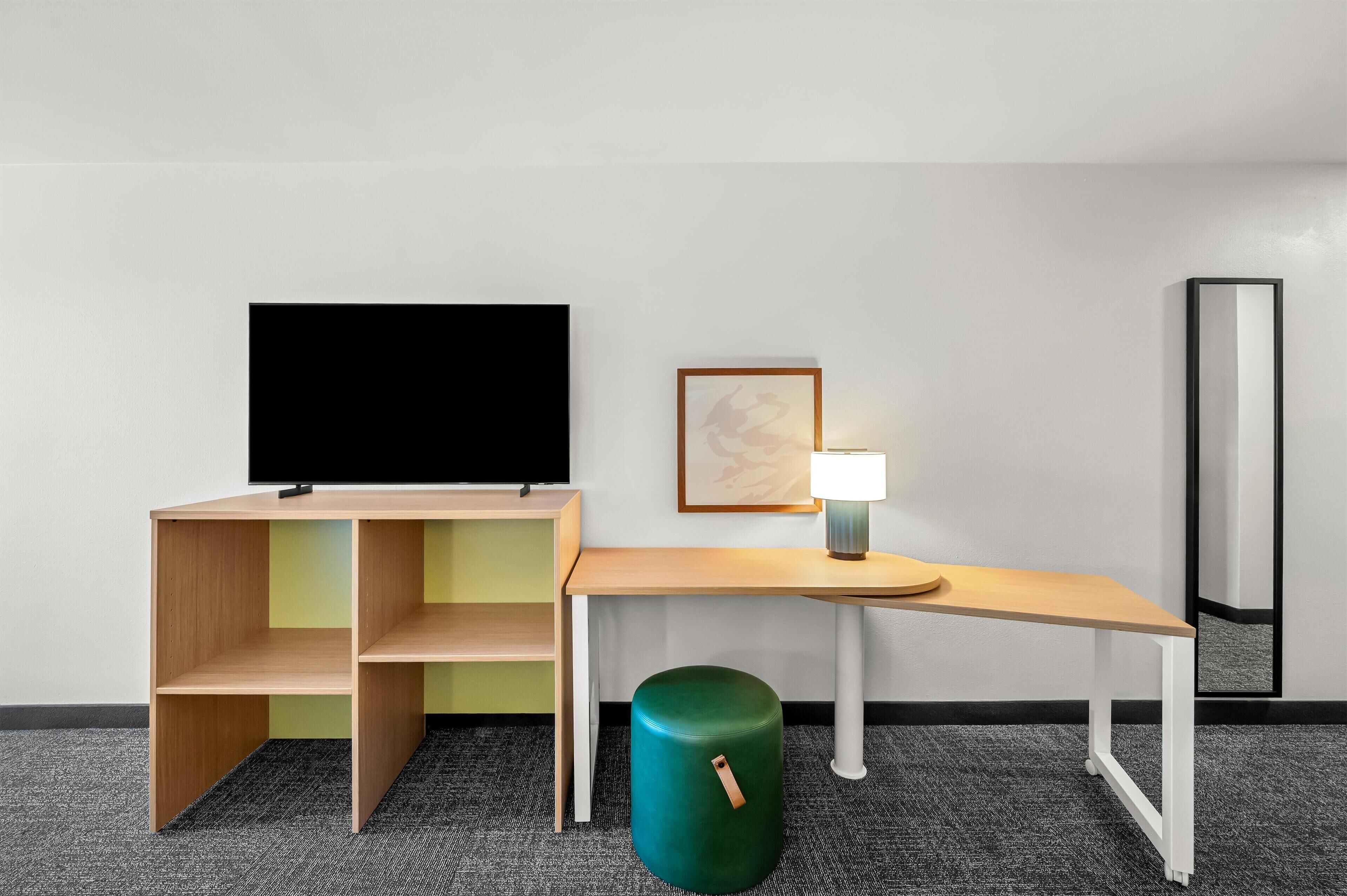 Una habitación de hotel con un escritorio de madera, un televisor de pantalla plana, una lámpara y un reposapiés verde.