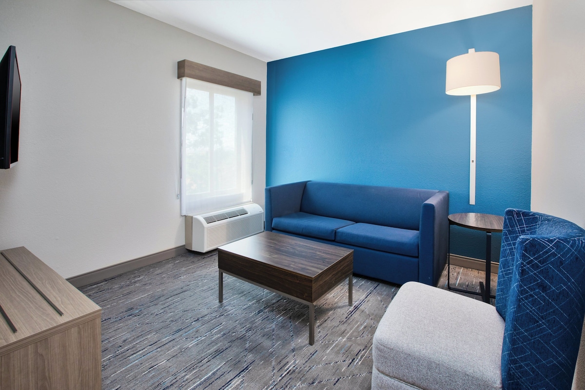 Quarto de hotel moderno com parede azul de destaque, sofá, mesa de centro, luminária e televisão.