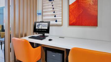 Een hotel business center met een computerbureau, twee oranje fauteuils, een printer en kunst aan de muur.