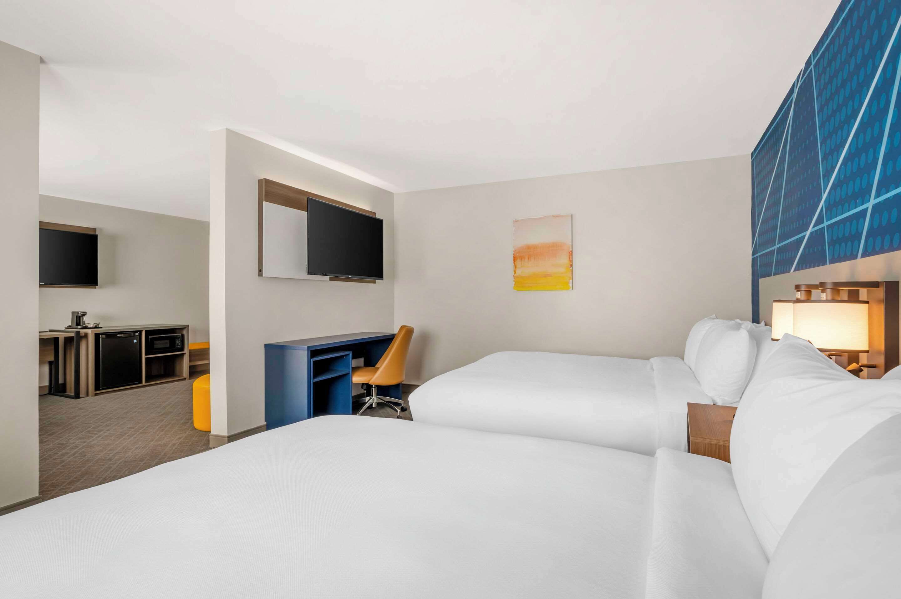 Habitación de hotel con dos camas, un escritorio con silla, televisor y un cuadro en la pared.
