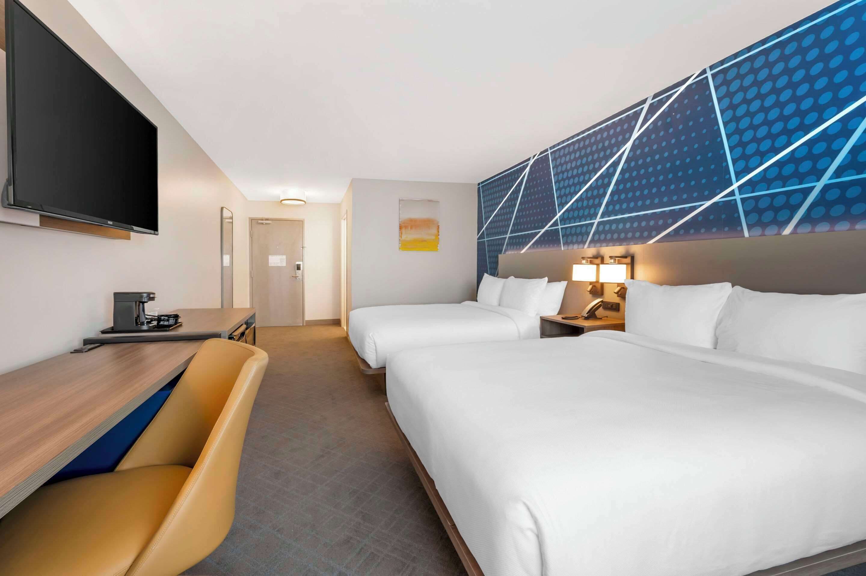 Une chambre d’hôtel avec deux lits, un bureau avec une chaise, une télévision à écran plat et un décor mural moderne.