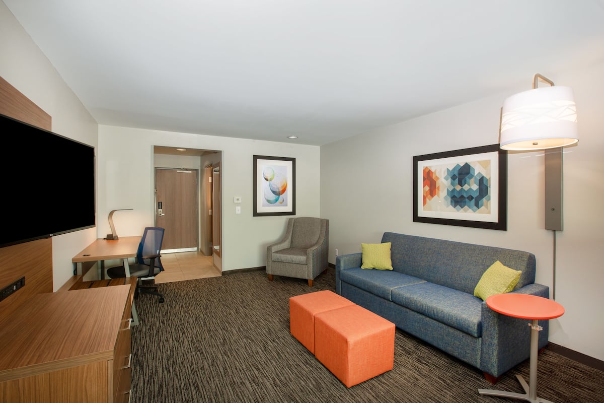 Ein modernes Hotelzimmer mit Flachbildfernseher, Schreibtisch mit Stuhl, blauem Sofa mit Kissen, orangefarbenem Hocker und Stehlampe.