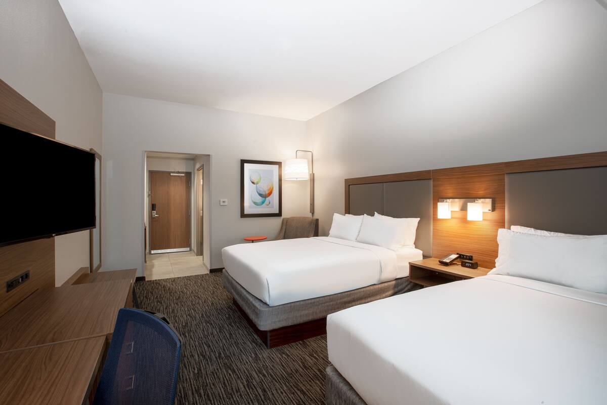 Ein Hotelzimmer mit zwei Betten, einem Schreibtisch, einem Sessel, einem Fernseher und einem gerahmten Bild an der Wand.