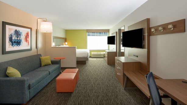 A hotel room with a sofa, ottoman, desk, TV, and a bed. - Holiday Inn Express Hotel & Suites Columbus by IHG (Columbus)