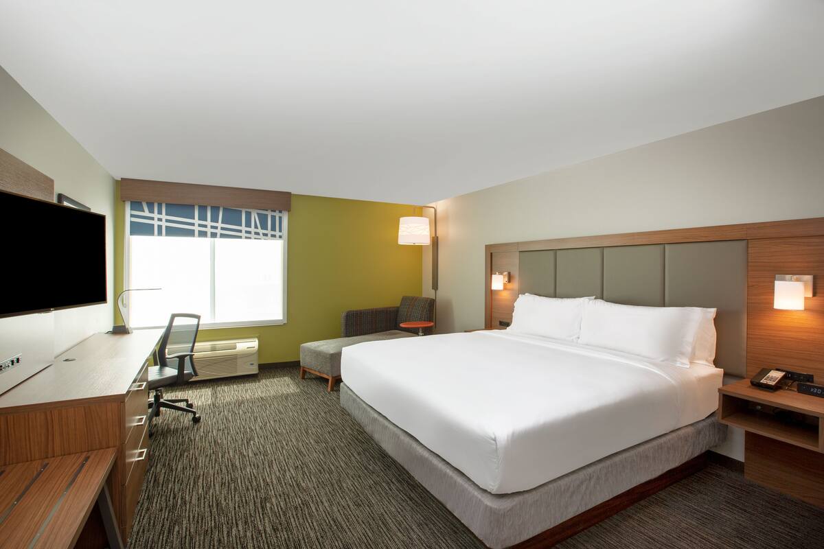 Ein Hotelzimmer mit einem großen Bett, einem Schreibtisch mit Computer, einem Sessel, einem kleinen Tisch und einem Fernseher.