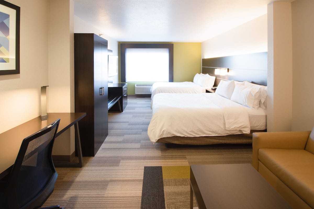 Habitación de hotel con cama, escritorio, silla, sofá y un amplio ventanal que proporciona luz natural.