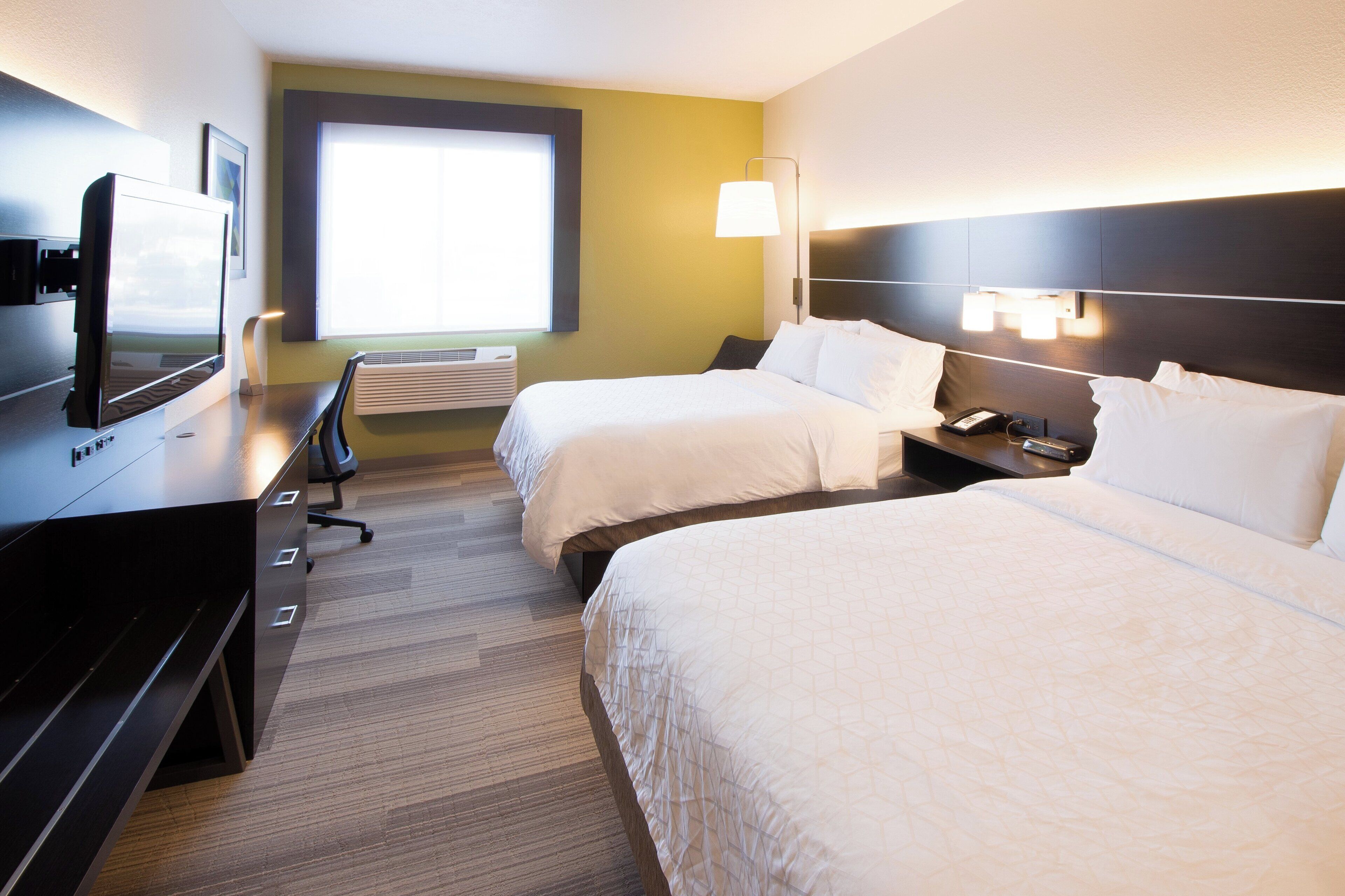 Ein Hotelzimmer mit zwei Betten, einem Schreibtisch mit Computer, einem Fernseher und einem Fenster mit Tageslicht.
