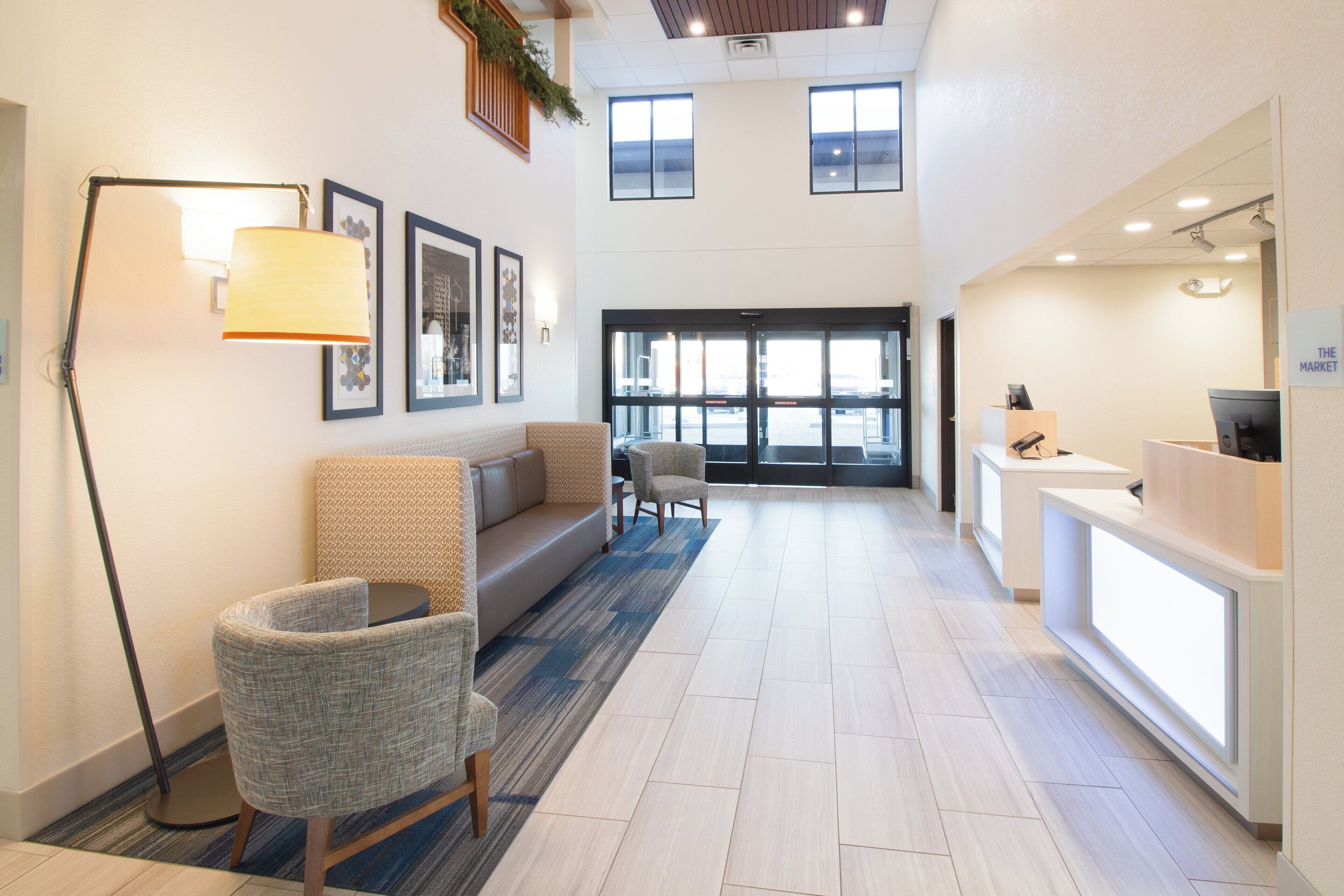 A hotel lobby with a reception desk, seating area with sofas and chairs, framed artwork on the walls, and large windows providing natural light.