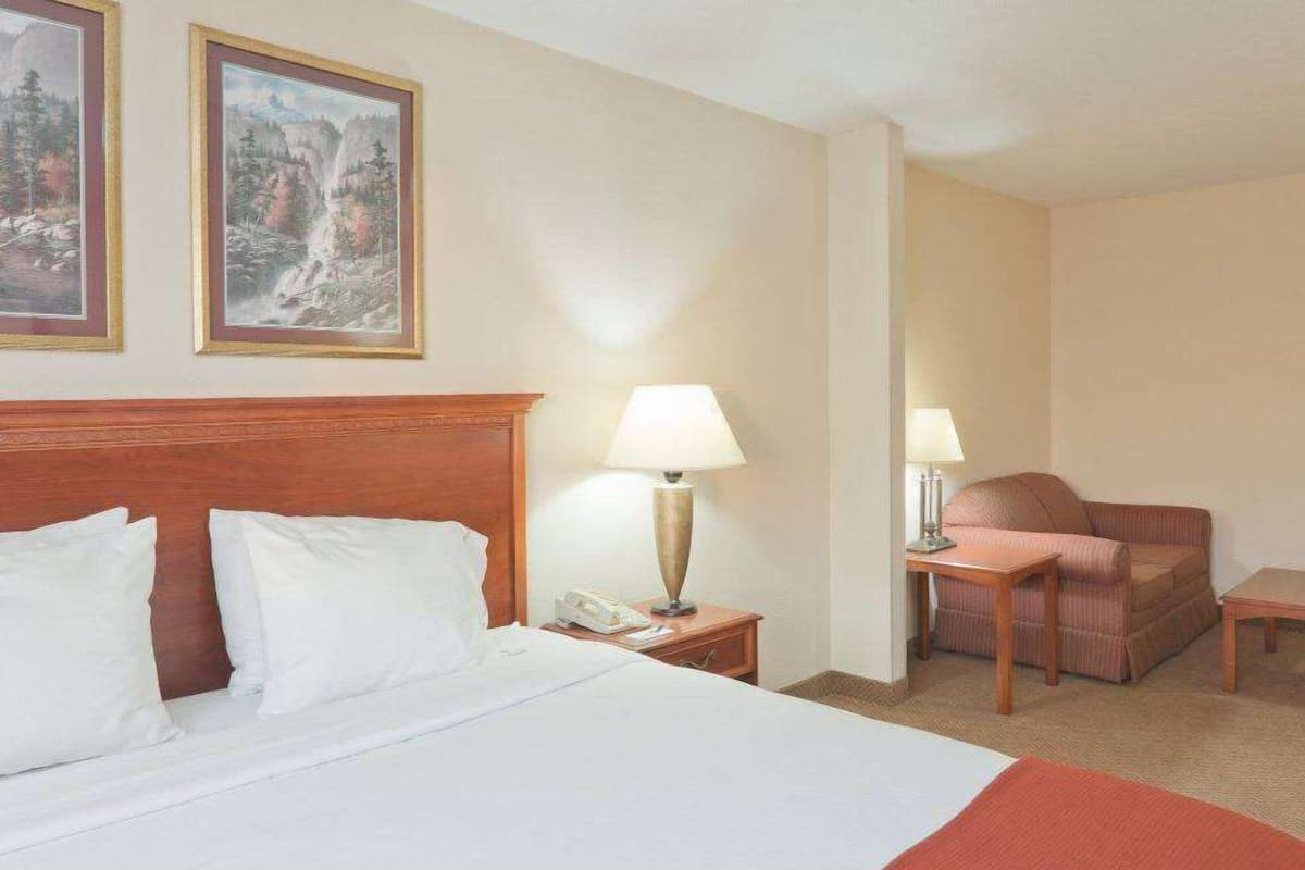 A hotel room with a bed, two bedside lamps, a chair, a small table, and two framed pictures on the wall.