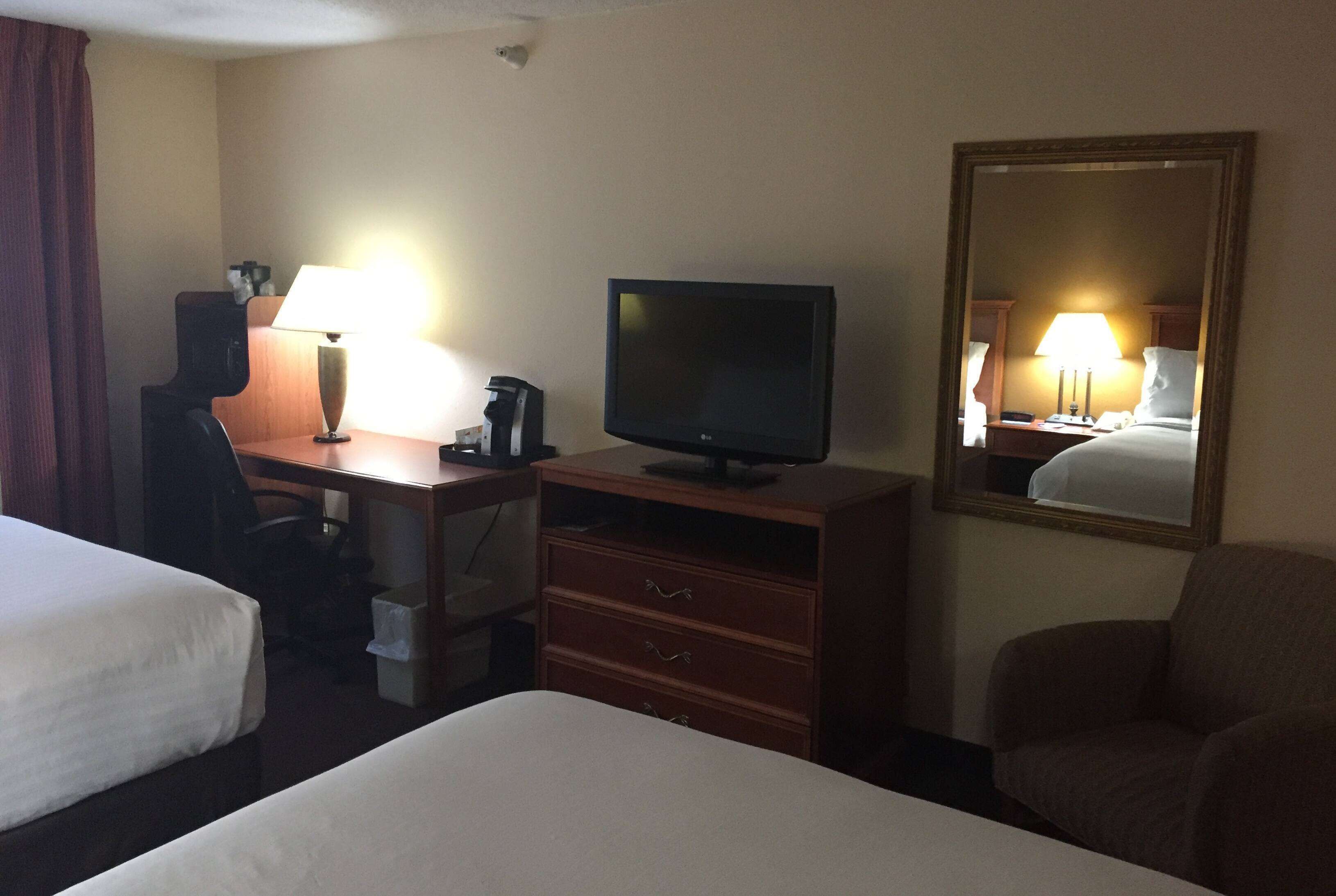 Habitación de hotel con dos camas, un escritorio con computadora y lámpara, un televisor, un espejo y una silla.