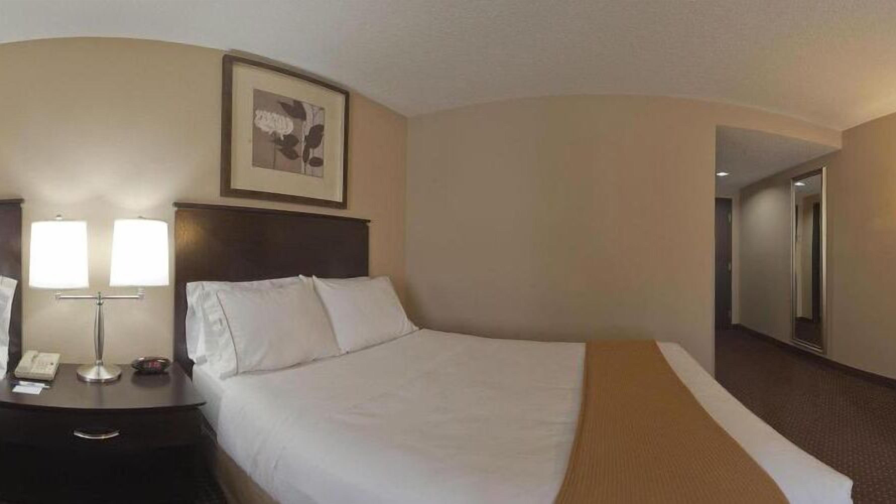 Une chambre d’hôtel avec un lit, une table de chevet avec une lampe, un tableau encadré au mur et une porte donnant sur une autre pièce.