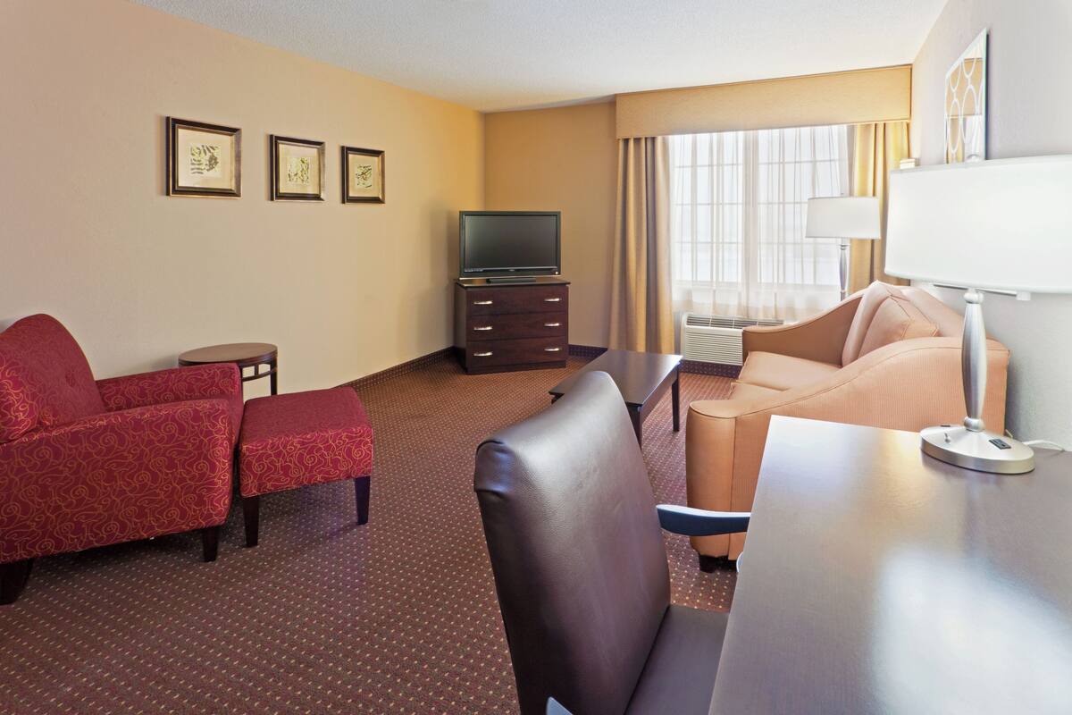 Ein Hotelzimmer mit einem roten Sofa, einem beigen Sofa, einem Fernseher auf einem Schrank, einer Lampe und einem Fenster mit Vorhängen.