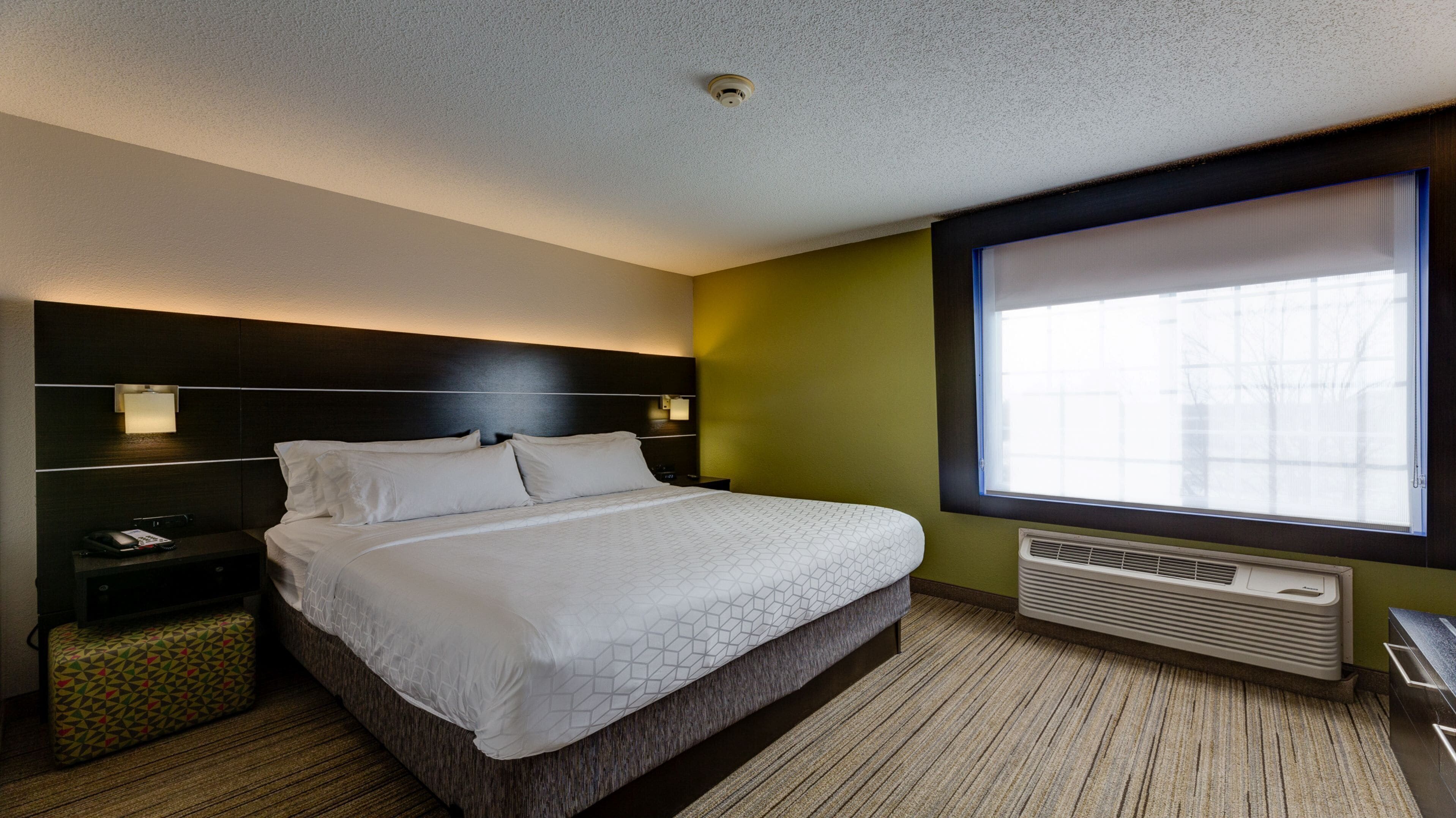 A hotel room with a bed, bedside tables, a telephone, a small bedside table with a patterned cushion, a window with blinds, and a wall-mounted air conditioner.