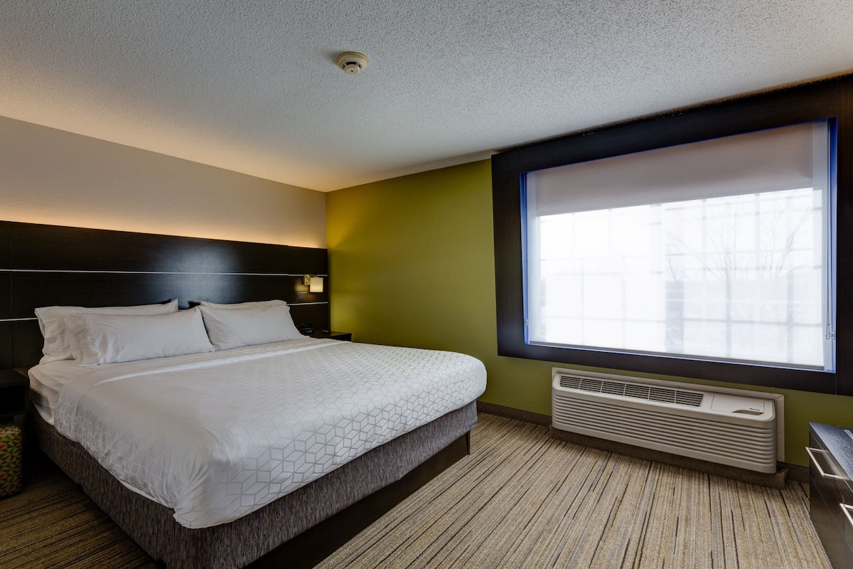 Ein Hotelzimmer mit Bett, Nachttischen, Telefon, einem kleinen Nachttisch mit gemustertem Kissen, einem Fenster mit Jalousien und einer Wand-Klimaanlage.